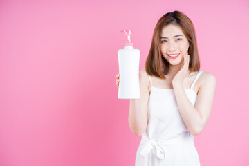 Image of young Asian woman holding shampoo bottle on pink background