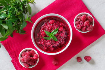 Delicious raspberry jam in a bowl, fresh berries, mint leaves on a pink napkin. Top view.