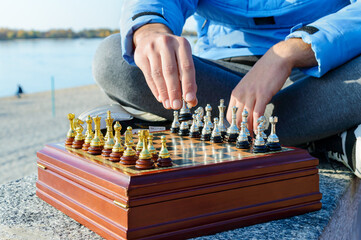 A young guy sits cross-legged and plays chess in the waterfront park. Close-up.