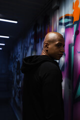 African american hooligan in hoodie looking away near graffiti on wall in garage