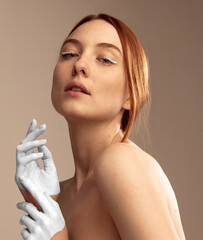 Portrait of tender red-haired woman with white painting on hands and white eyeliner posing isolated on beige background