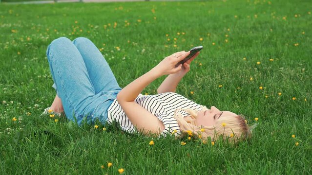 blond Caucasian girl lying on the grass and using a mobile phone. outdoor full shot. High quality 4k footage
