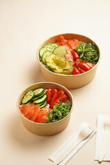 Trendy wholesome dish poke or buddha bowl - rice, wakame seaweed, tomatoes, cucumber, and red fish salmon - in a recycled round carton with sustainable fork, takeaway food concept on beige background