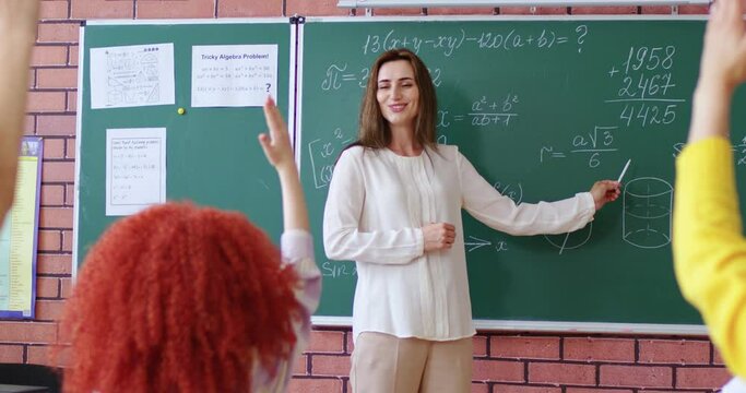 Unrecognizable schoolgirl raising her hand and answering question asked by female teacher in math class. Smiling tutor standing near blackboard and asking math rules.