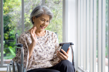 Fototapeta na wymiar Asian gray hair old woman waving her hand via remote communication through mobile phone while sitting on wheelchair in front of corridor with glass wall.
