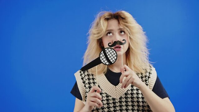 Funny blond woman with fake mustache and paper microphone pretending to sing. Isolated on blue background. Blue November concept