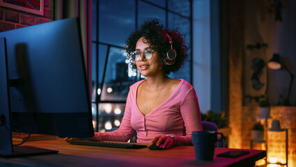 Fototapeta na wymiar Young Beautiful Black Woman in Headphones Using Computer in Stylish Loft Apartment in the Evening. Creative Female Smiling, Browsing Videos on Social Media. Urban City View from Big Window.