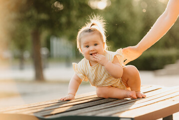 A female toddler at the bench near a hand of a mom in the park in the sun lights.