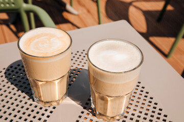 Two large transparent glasses of cappuccino with coffee art on a beige table in bright sunlight. Coffee in a cafe
