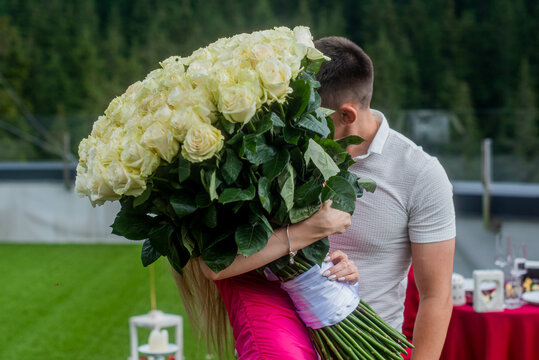 The Bride In A Pink Dress Holds A Large Bouquet Of White Roses. Kiss Of Newlyweds Covered With A Bouquet On The Background Of An Outdoor Dinner