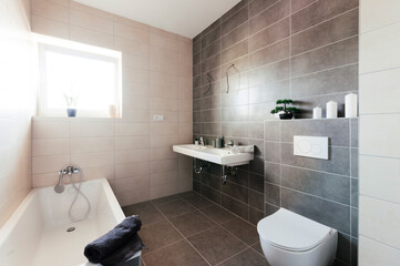Unfinished modern bathroom in the apartment. There is no mirror on the wall, instead of lights there are cables hanging from the walls. The sanitary ware is white and tiling is in brown-beige shade.