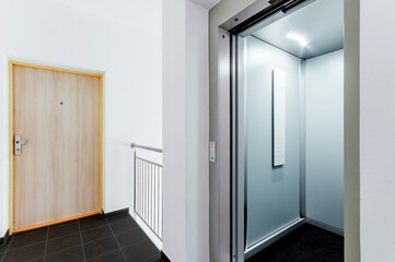 Hallway in building with open elevator door and entrance wooden door with security lock and optical peephole. The elevator is modern with a touch panel and digital display.