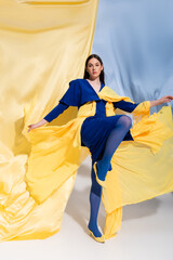 full length of young ukrainian woman in stunning color block clothing posing near blue and yellow fabric.