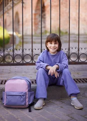 Abwaschbare Fototapete Süße Monster Fröhliches junges Mädchen, das in der Stadt in der Nähe ihres Rucksacks auf dem Boden sitzt