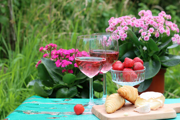 Two glasses of rose wine, pink flowers, strawberry, brie cheese and fresh croissants on a table. Picnic in the garden. Summer food on a table. 