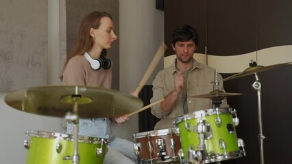 Young woman takes drum lesson from male teacher in professionally equipped music studio. Woman energetically plays drums and cymbals man watches performance