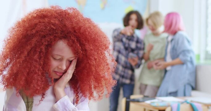 Crying girl with red curly hairstyle suffering from bullying by peers at school. Group of schoolchildren scoffing at their classmate on blur background.