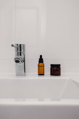 Beauty cosmetic product bottle in brown glass on a white sink in bathroom background with copy space. Eco friendly sustainable skin care. Minimal home interior. Morning beauty routine.