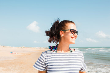 Portrait close-up happy brunette woman in a striped dress wearing sunglasses stands by the sea, looks at the waves, laughs and smiles. Tourism, beach holidays, breeze, journeys, travel