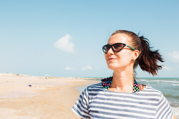 Portrait close-up happy brunette woman in a striped dress wearing sunglasses stands by the sea, looks at the waves, laughs and smiles. Tourism, beach holidays, breeze, journeys, travel