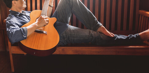 An Asian man is lying on a wooden sofa playing the guitar in a relaxed mood.on vacation at home