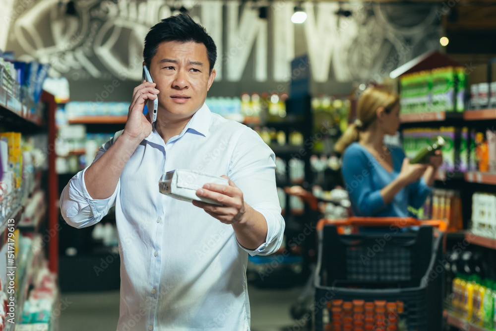 Wall mural An Asian man cannot choose a product in a supermarket, has difficulty choosing, calls his wife for advice