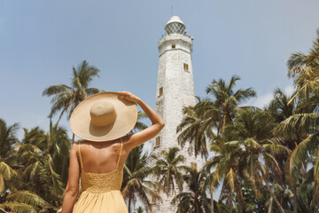Back view of romantic woman traveler in dress and straw hat visiting famous landmarks of Sri Lanka country standing and looking on Dondra lighthouse. High quality photo. - obrazy, fototapety, plakaty