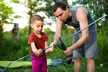 Father showing to son how to assemble rack for tent in hike on summer sunset outdoors