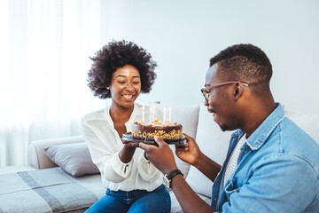 Multiethnic couple sitting at table and celebrating birthday at home in living room. Young african girl surprised on seeing birthday cake with her boyfriend. 