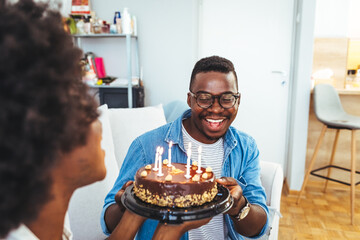 Happy millennial couple celebrating birthday with festive cake at home, full length. Cheerful African American  guy greeting excited girlfriend with b-day, enjoying holiday in living room