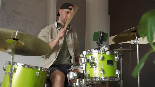 Young man greets followers recording online drum lesson on mobile phone and plays drums in equipped music studio. Guy explains nuances of drums playing technique