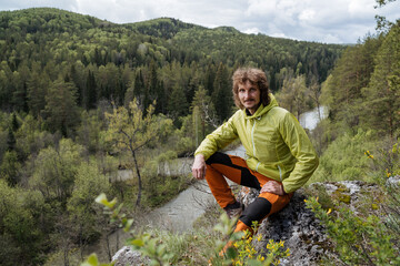 A tourist sits on a stone against the background of nature, a guy in a jacket travels through the taiga, a man stands on the edge of a cliff, the river flows below, a green forest. © Aleksey