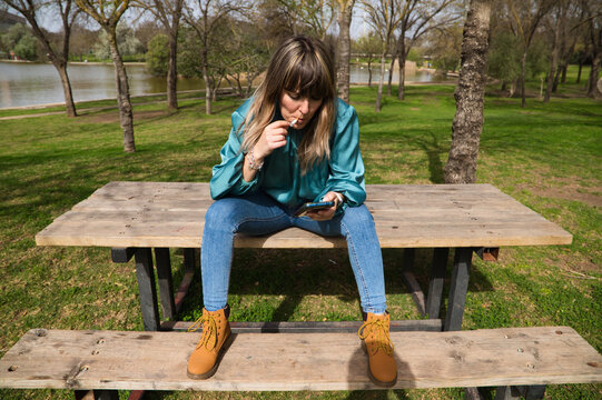 A young, pretty, blonde woman is sitting at a wooden table in the park, looking at her mobile phone and smoking a cigarette. In the background you can see the river on a sunny day. Stop smoking