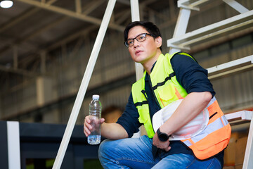 Time for a tea break concept. Asian male warehouse Industrial, factory and energy engineer Specialist drinking water sitting on stair at warehouse office factory workplace.