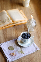 Vintage porcelain cup filled with fresh blueberries, pressed flowers, open book, reading glasses and lit candle on the table. Hygge at home. Selective focus.