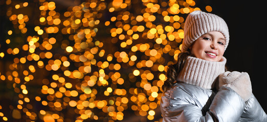 Girl and Christmas lights. Smiling little girl in a bright hat, scarf and jacket at night with blurry lights. Holidays theme. Banner