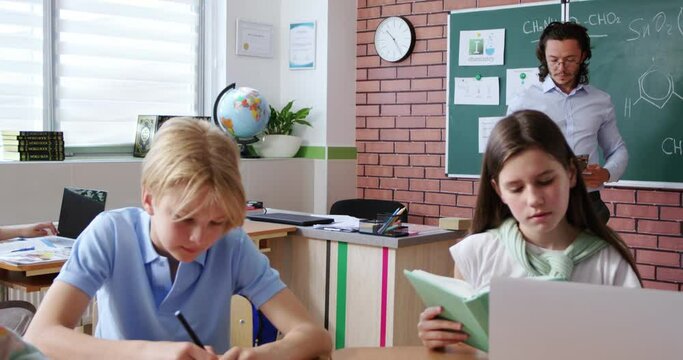 Enthusiastic caucasian teacher showing digital tablet to male and female pupils sitting together at desk. Smart kids learning new stuff at modern school with help of different gadgets.