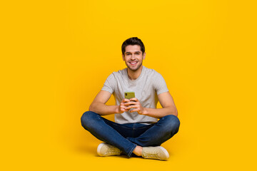 Full size photo of funny brunet young guy sit hold telephone wear t-shirt jeans sneakers isolated on yellow background