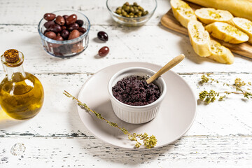 Olive pate in ramequin bowl and basic ingredients - olive oil, olives and capers on white wooden background