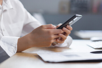Close-up photo. The hands of a young woman in a white shirt are holding a mobile phone, typing a message at the table.