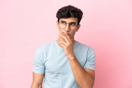 Young Argentinian man isolated on pink background With glasses and having doubts