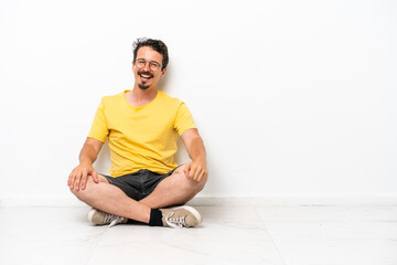 Fototapeta premium Young caucasian man sitting on the floor isolated on white background with glasses and happy