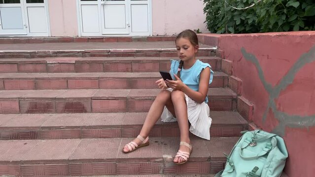 A little girl is playing a tablet while sitting alone in the street. the girl is doing lessons in nature, it's time to go to school, the girl maintains her blog, communicates with subscribers or frien
