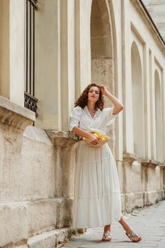 Fototapeta Beautiful curly brunette woman wearing trendy summer white linen shirt, long skirt, sandals, holding bowl with lemons, posing in street of European city. Full-length outdoor portrait
