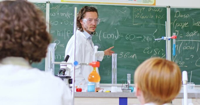 Male teacher in protective glasses and white lab coat standing near blackboard with writing chemical formulas and talking to children. Pupils sitting at desk during chemistry lesson.