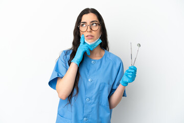Young woman dentist holding tools isolated on white background having doubts while looking up