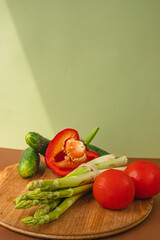Vegetables lie on a wooden board: tomatoes, asparagus, cucumbers, red bell peppers. brown, light green background. place for text.