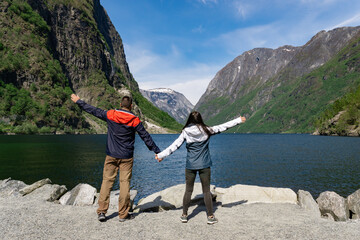 Fototapeta na wymiar Unrecognizable tourist couple with their arms up with their backs turned, at the foot of the fjord and surrounded by high mountains in Gudvangen - Norway.
