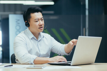 Fototapeta na wymiar A young handsome Asian businessman in headphones with a microphone sits at a laptop in the office, conducts an online conference, a remote meeting with clients, partners. Tells, explains, smiles