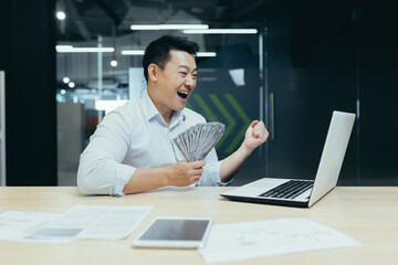 Fototapeta na wymiar A happy and smiling young Asian businessman concluded a profitable financial deal with clients and partners. He sits in the office at a table with a laptop, shows and holds g of money in his hands.